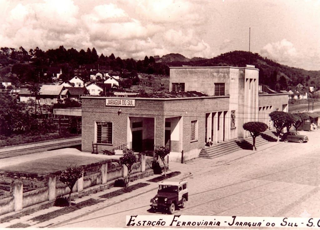 Fachada da Estação Ferroviária de Jaraguá do Sul vista da rua, com placa identificadora no alto do prédio e um jeep antigo no primeiro plano, registro do período em que a estação era o centro social da cidade