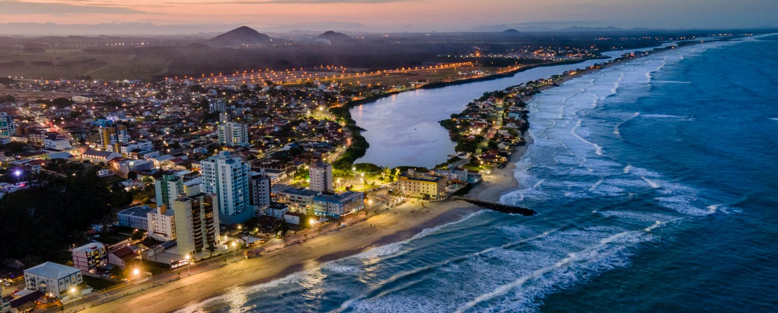 Vista aérea de Barra Velha ao entardecer, com a orla, o mar e a foz do rio encontrando a praia