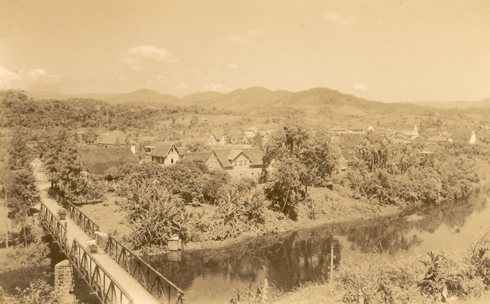 Foto antiga em sépia mostra Jaraguá do Sul no passado, com ponte sobre o rio, casario e os morros do vale ao fundo