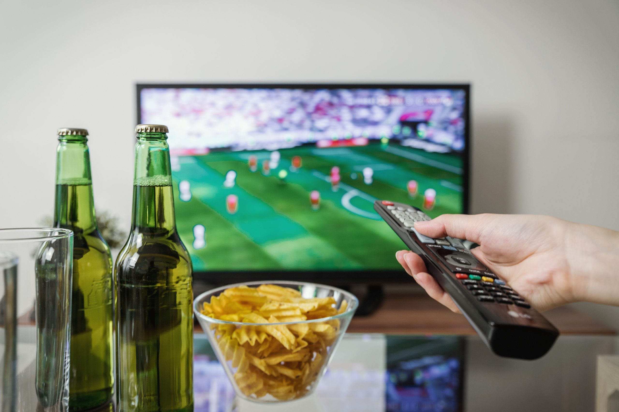 Torcedor assiste a uma partida de futebol na televisão enquanto segura o controle remoto, com bebidas e petiscos sobre a mesa.