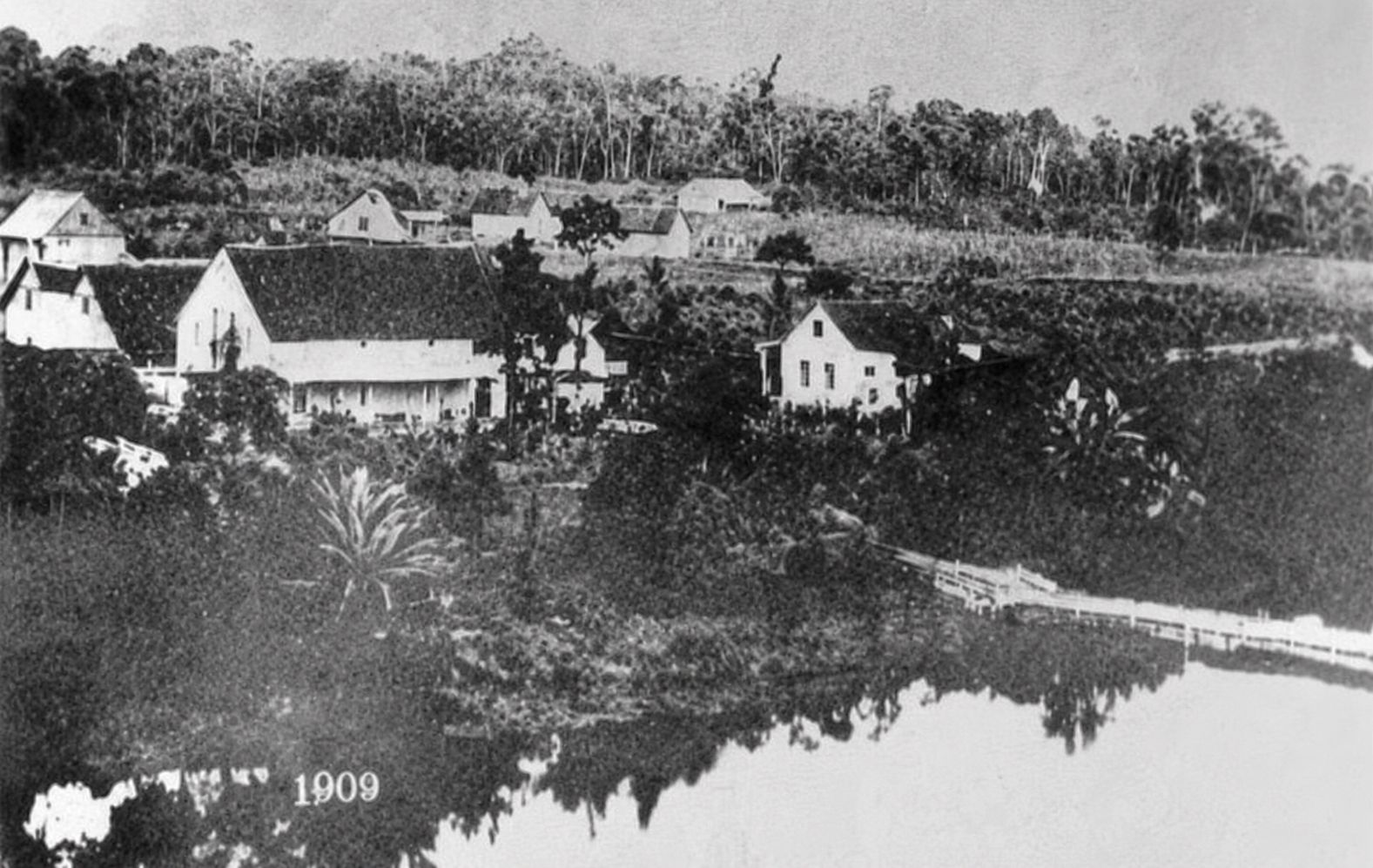 Povoado de Jaraguá do Sul em 1909, com casas na encosta e a primeira ponte de madeira sobre o Rio Itapocu à direita